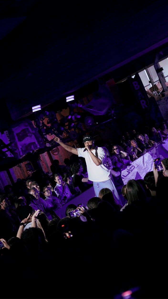 Le chanteur Disiz se produit sur scène micro à la main et bras levé face au public lors de l'événement de lancement de la CUPRA Raval à Paris. La scène est plongée dans une ambiance lumineuse violette où de nombreux spectateurs le filment avec leur smartphone.