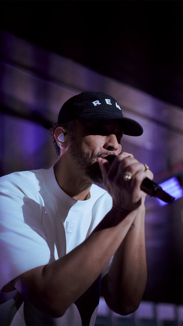 Gros plan du chanteur Disiz se produisant micro à la main lors du lancement de la CUPRA Raval à Paris, portant une casquette noire et un t-shirt blanc dans une ambiance lumineuse violette.