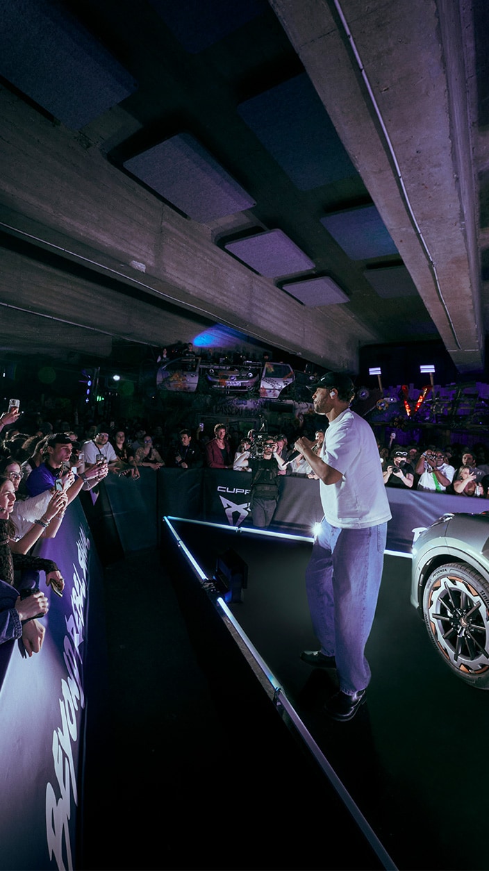 Le chanteur Disiz se produit sur une scène en V aux bordures LED bleues, face à une foule massée derrière des barrières CUPRA. Portant un t-shirt blanc et un jean ample, il chante sous un plafond en béton brut dans une ambiance lumineuse violette, avec une partie de voiture visible à droite.