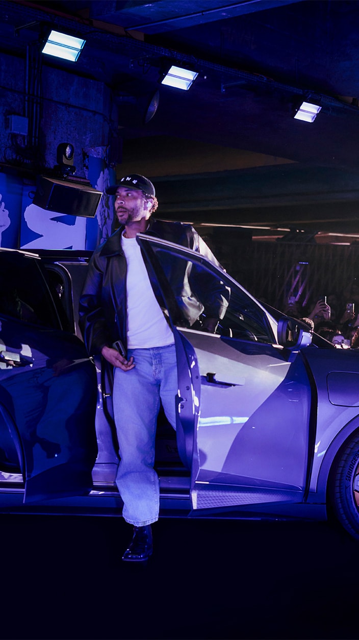 Le chanteur Disiz, vêtu d'un blouson en cuir noir, d'un t-shirt blanc et d'un jean ample, descend d'une CUPRA Raval dans un décor industriel. La scène est baignée d'une lumière bleue intense avec une foule visible à l'arrière-plan.