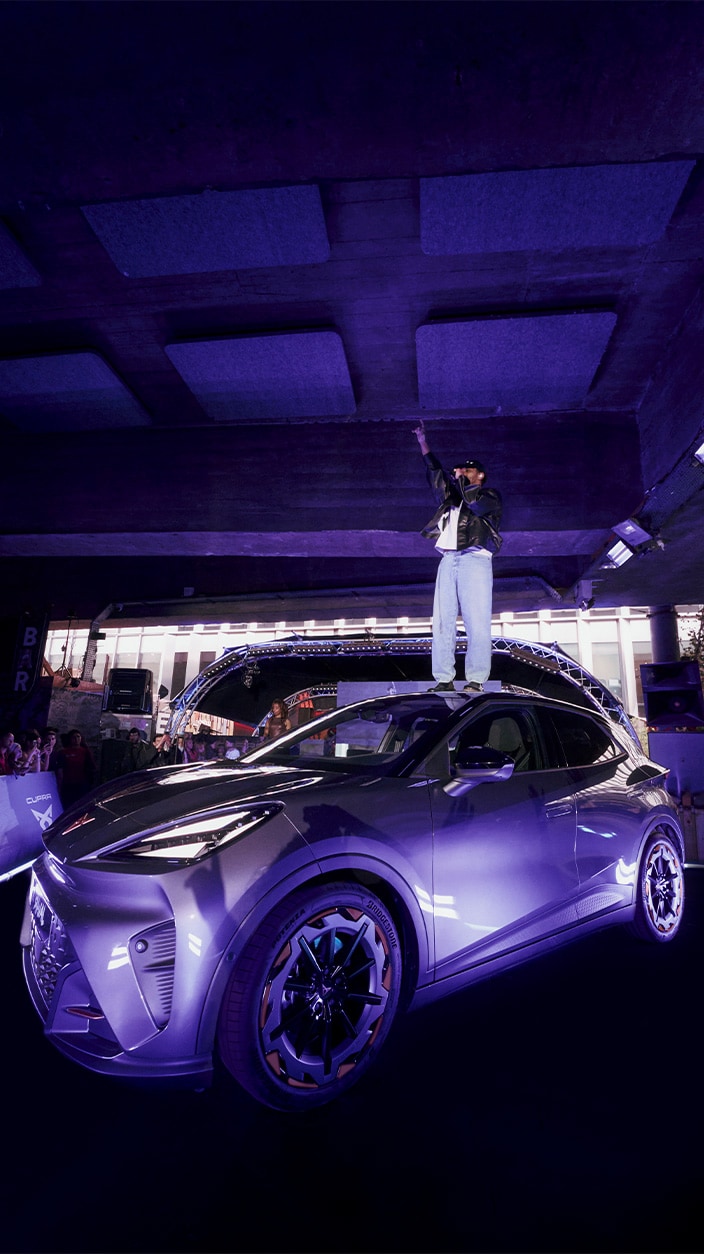 Vue en contre-plongée de la CUPRA Raval lors de son événement de lancement à Paris, éclairée par des lumières violettes sous une structure en béton. Le chanteur Disiz se tient debout sur le toit du véhicule, levant un bras vers le haut face à la foule présente en arrière-plan.