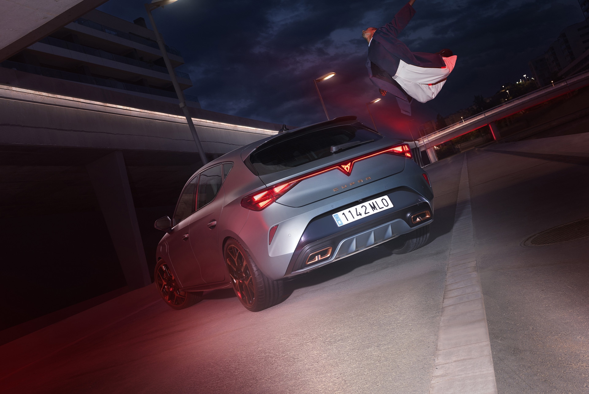 Technologie de la nouvelle Cupra Leon 2025 hybride, éclairage LED arrière, véhicule PHEV garé sur le ciment, structure avec des bords lumineux. Le véhicule est présenté dans une finition Gris mat avec des jantes forgées en Cuivre et Noir.