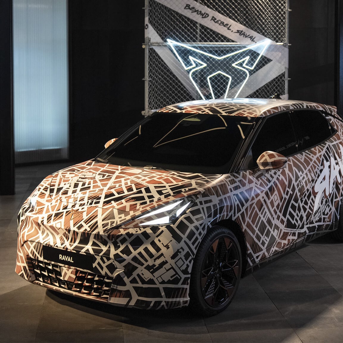 La CUPRA Raval, recouverte d'un habillage camouflage marron et blanc représentant un plan de ville, est exposée à l'intérieur. Derrière la voiture se trouve un panneau de clôture grillagée avec le logo CUPRA blanc lumineux et le texte « BEYOND REBEL, RAVAL. ».