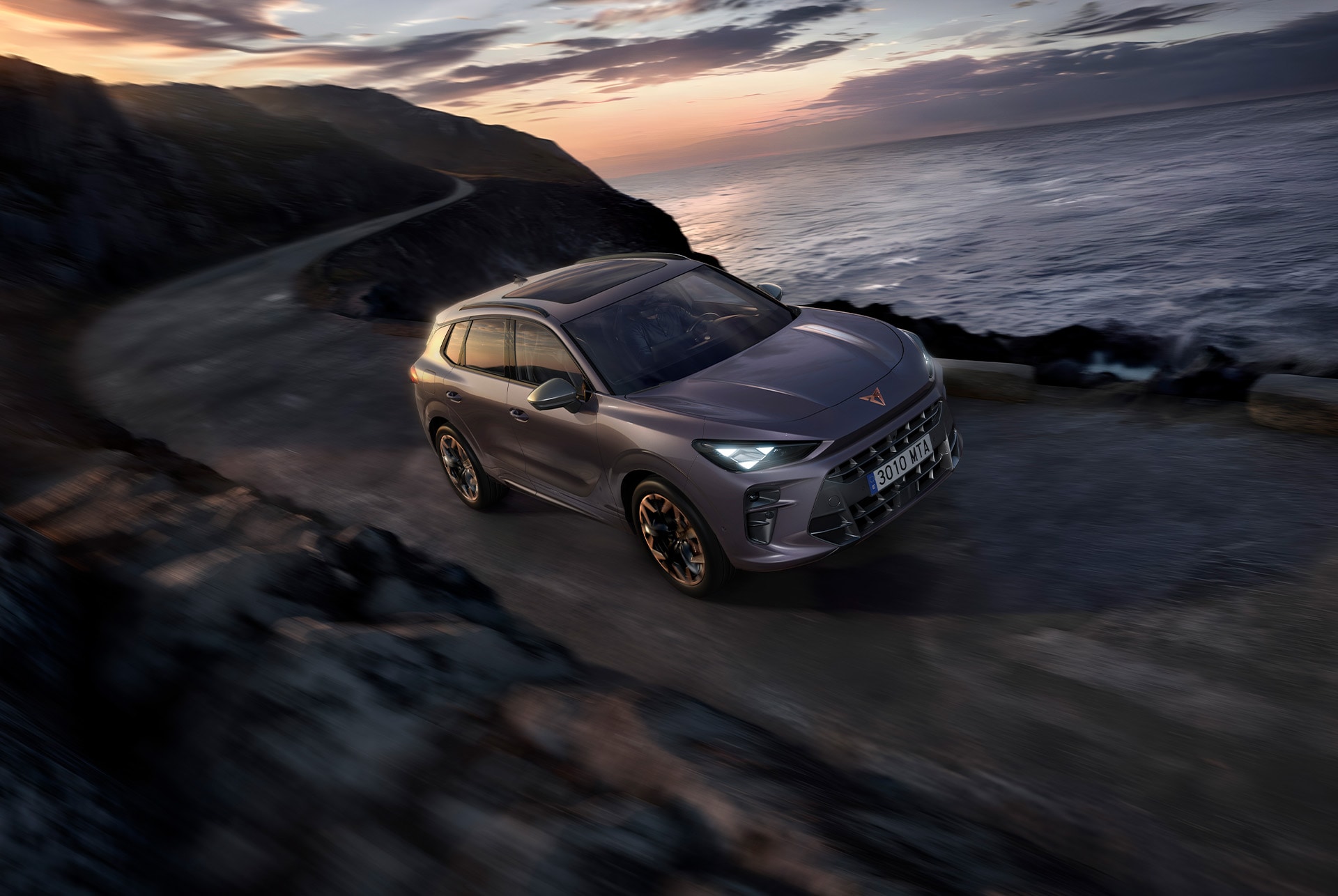 CUPRA Terramar avec le réglage adaptatif du châssis (dcc), sur une route sinueuse et mouillée devant le coucher du soleil en bord de mer.