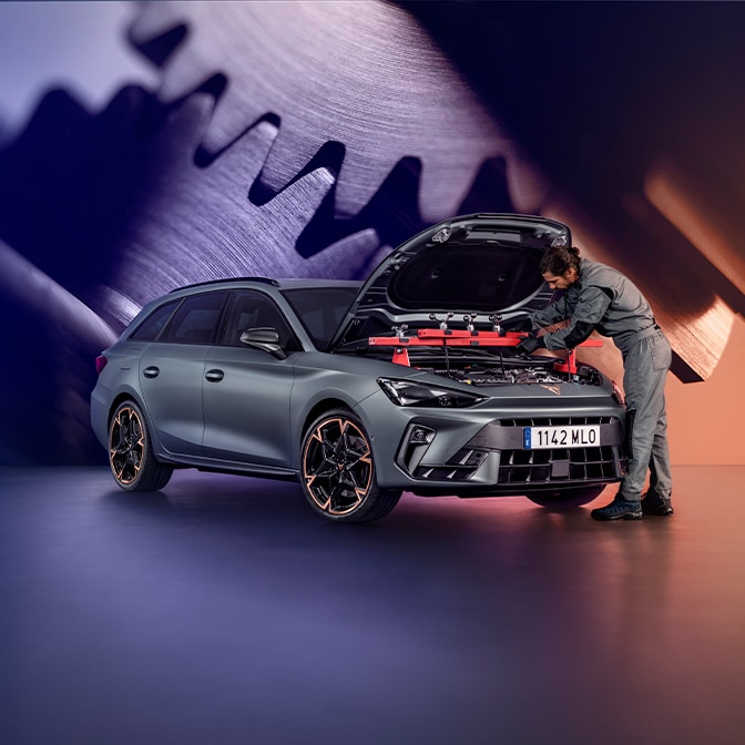 A technician in a grey jumpsuit inspecting the engine bay of a CUPRA Terramar in a studio; the background features a large screen displaying a close-up of metallic gears.