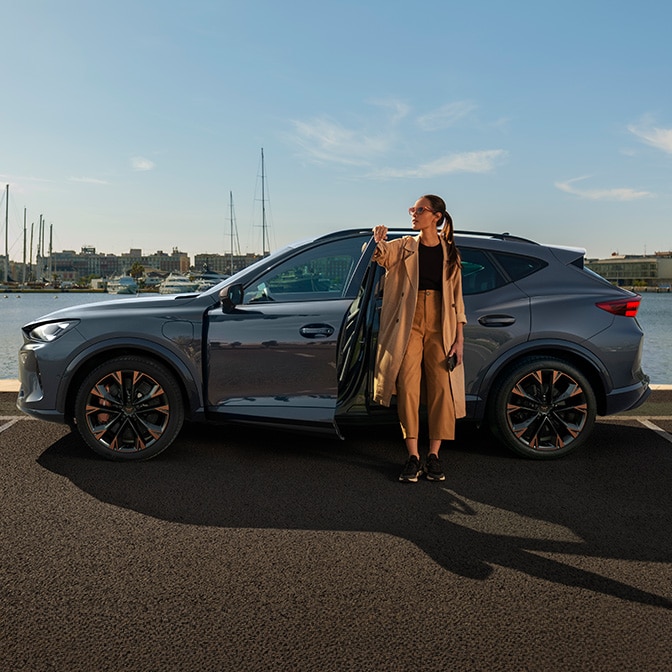 A woman leaning against a grey CUPRA Leon and looking at her phone while the car is plugged into a wall-mounted charger outside a modern, minimalist wood and concrete house.