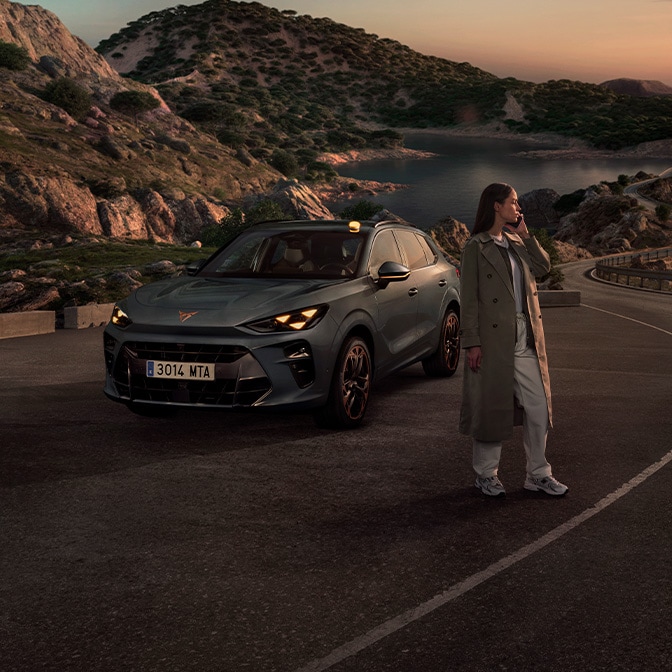 A wide-angle landscape shot at dusk showing a woman in a trench coat standing on a winding coastal road next to a CUPRA Terramar, with rocky hills and a calm sea in the background.