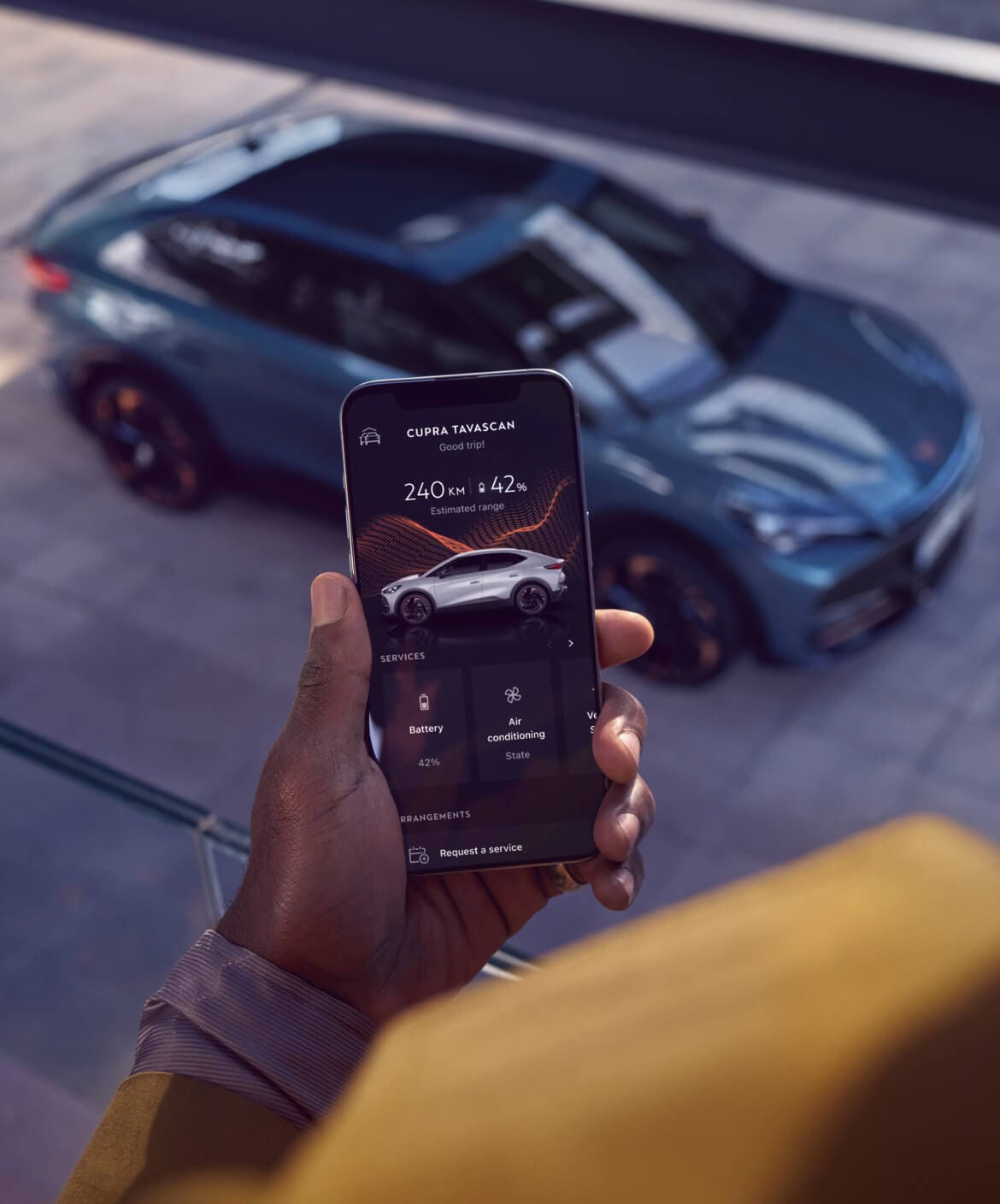 Une personne tenant un smartphone sur lequel est affichée l’application My CUPRA. CUPRA Tavascan VZ en arrière-plan, idéal pour la location de voitures d’entreprise ou le leasing.