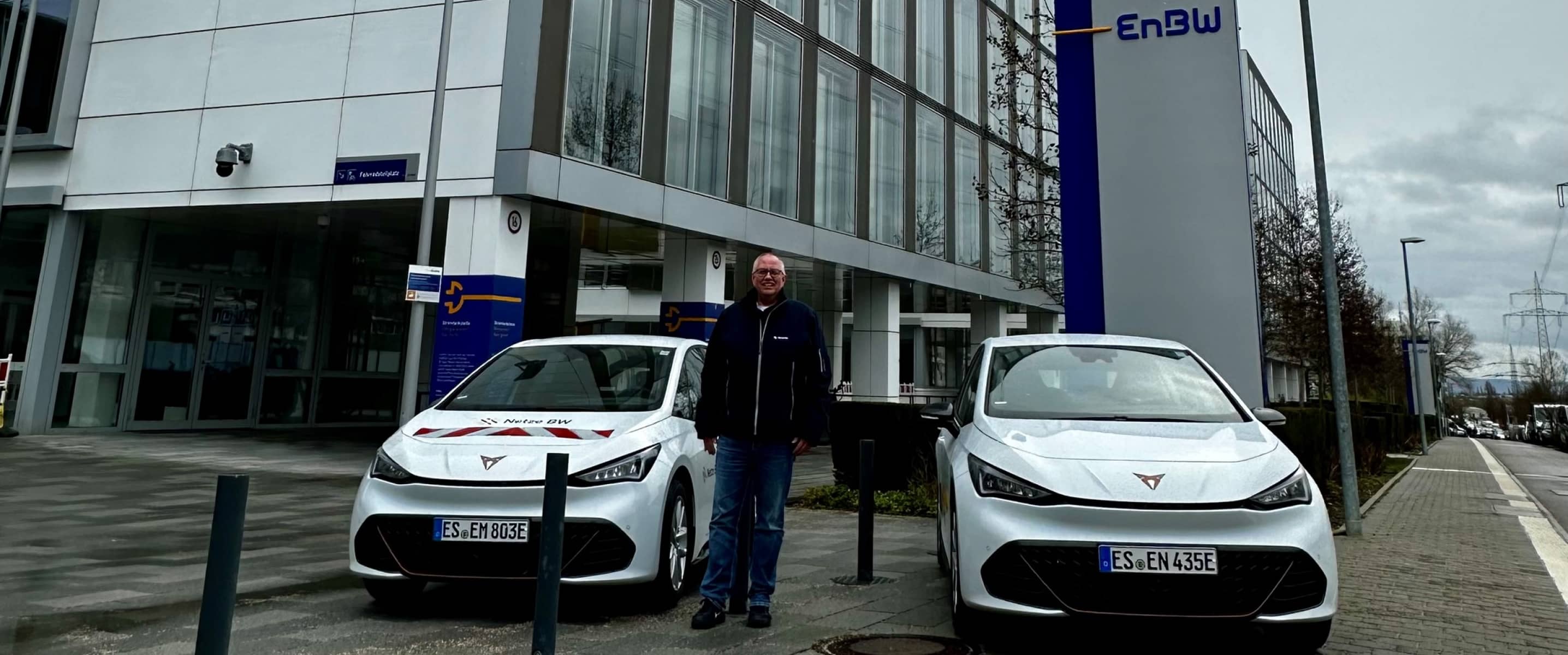 Deux véhicules cupra born blancs garés devant un immeuble de bureaux enbw, avec un gestionnaire de flotte se tenant entre les voitures par une journée nuageuse.