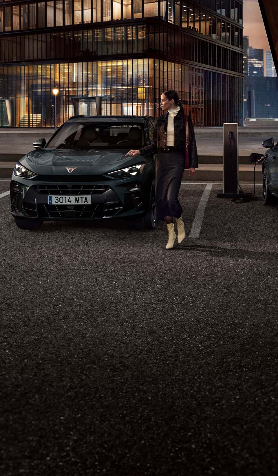 Une femme marche entre deux voitures CUPRA stationnées, dont l’une est raccordée à une borne de recharge publique, avec des immeubles en arrière-plan.