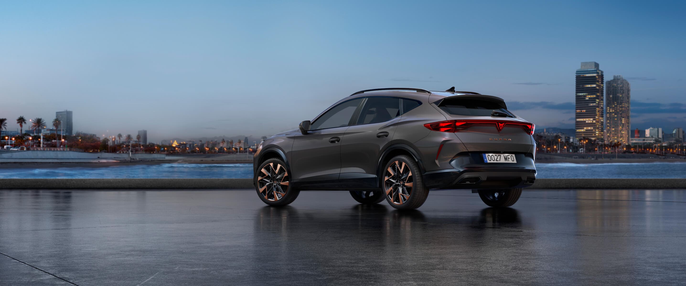 Qu’est-ce qu’un véhicule hybride léger, une Cupra Formentor en gris graphite, vue arrière, garée au bord de l’eau au coucher du soleil avec la ligne d’horizon de la ville.