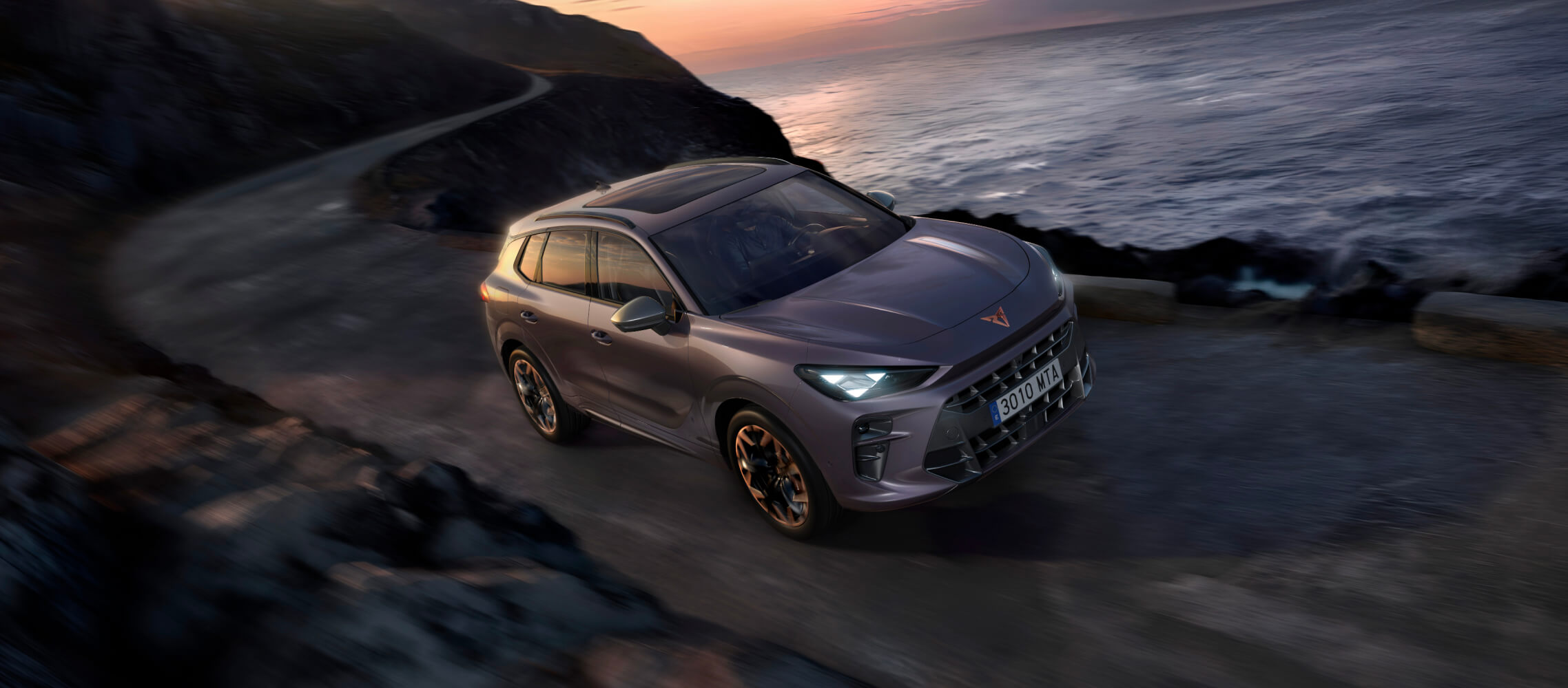 Une voiture électrique Terramar, avançant sur une route rocheuse, avec la mer d’un côté et un coucher de soleil haut en couleur en arrière-plan.