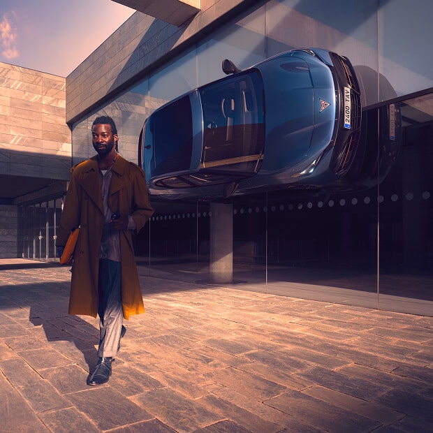 A person in a long coat on a patterned stone ground, walking away from a modern building with a CUPRA vehicle mounted vertically on its glass facade.